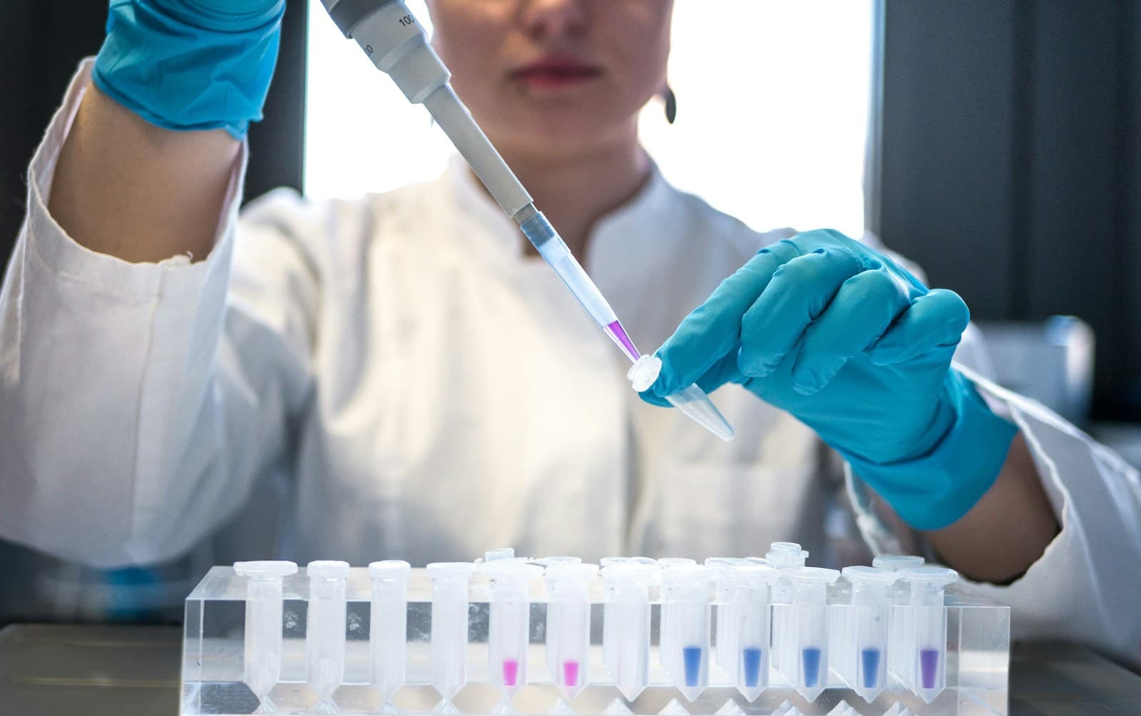 A scientist in a white coat and gloves pipetting into a rack of small tubes