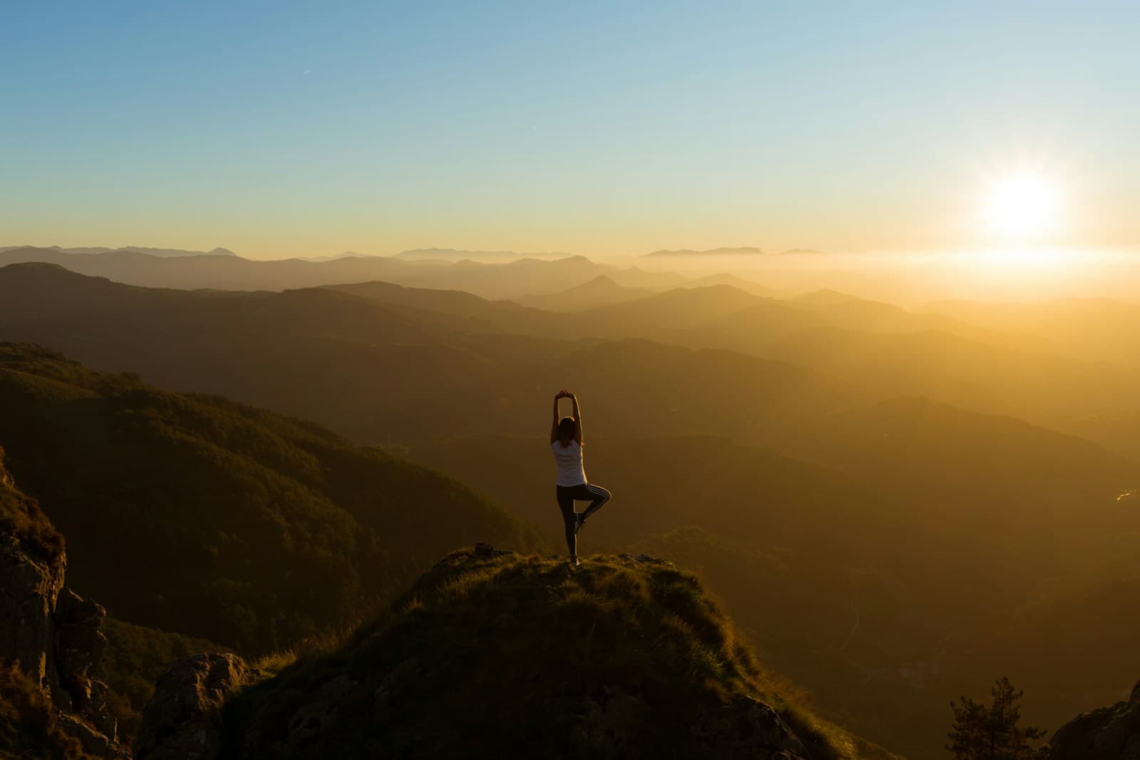 A person holding a yoga tree pose on a mountaintop at sunrise, above misty hills